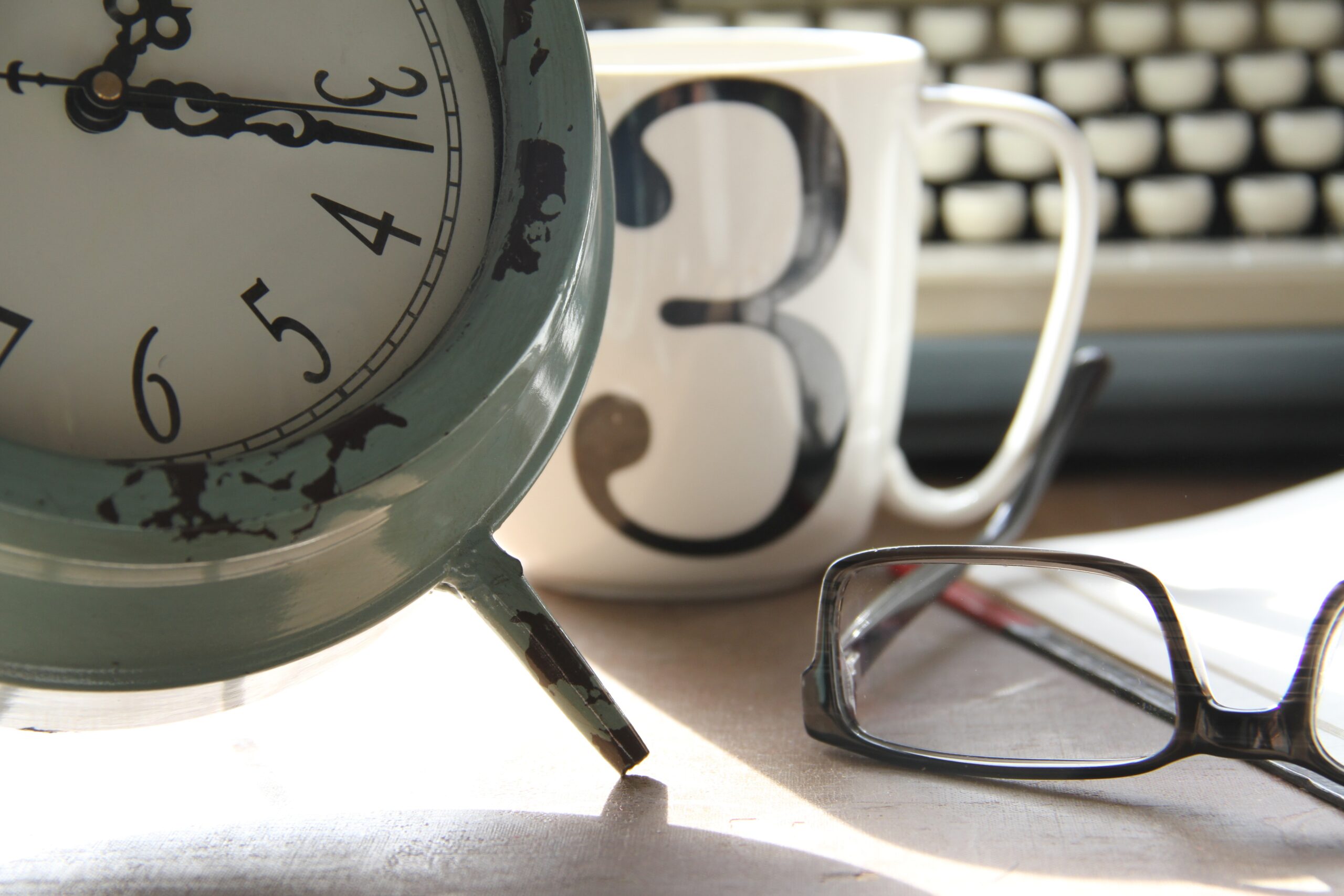 Scrivania con una sveglia, una tazza, occhiali e una tastiera di pc: nella procrastinazione si rimanda il compito importante per attività secondarie
